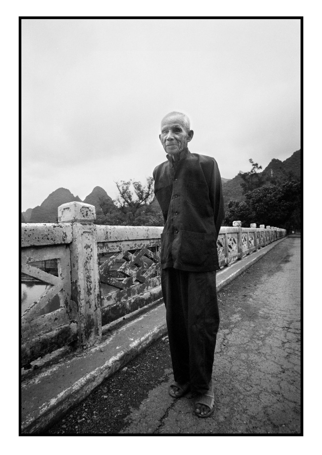 Photo, Voyage, Village, Extérieur, Nature, Montagne, Arbre, Palmier, Maison, Personnage, Homme, Personne âgée, Portrait, Rides, Asiatique, De pied, Tenue traditionnelle, Regard, Pont, Balustrade, Rivière, Eau, Reportage, Noir et blanc, n&b, nb, Chine, Asie